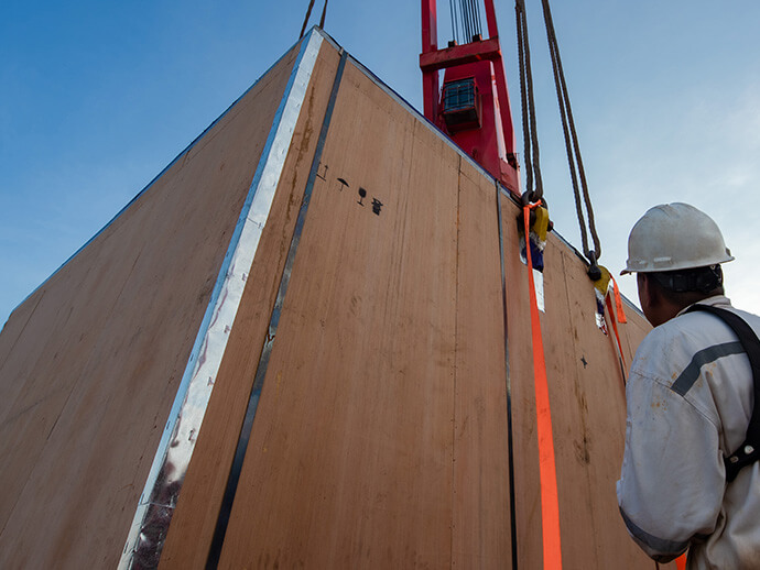 Man in hardhat and coveralls guides a large box as it is unloaded by crane